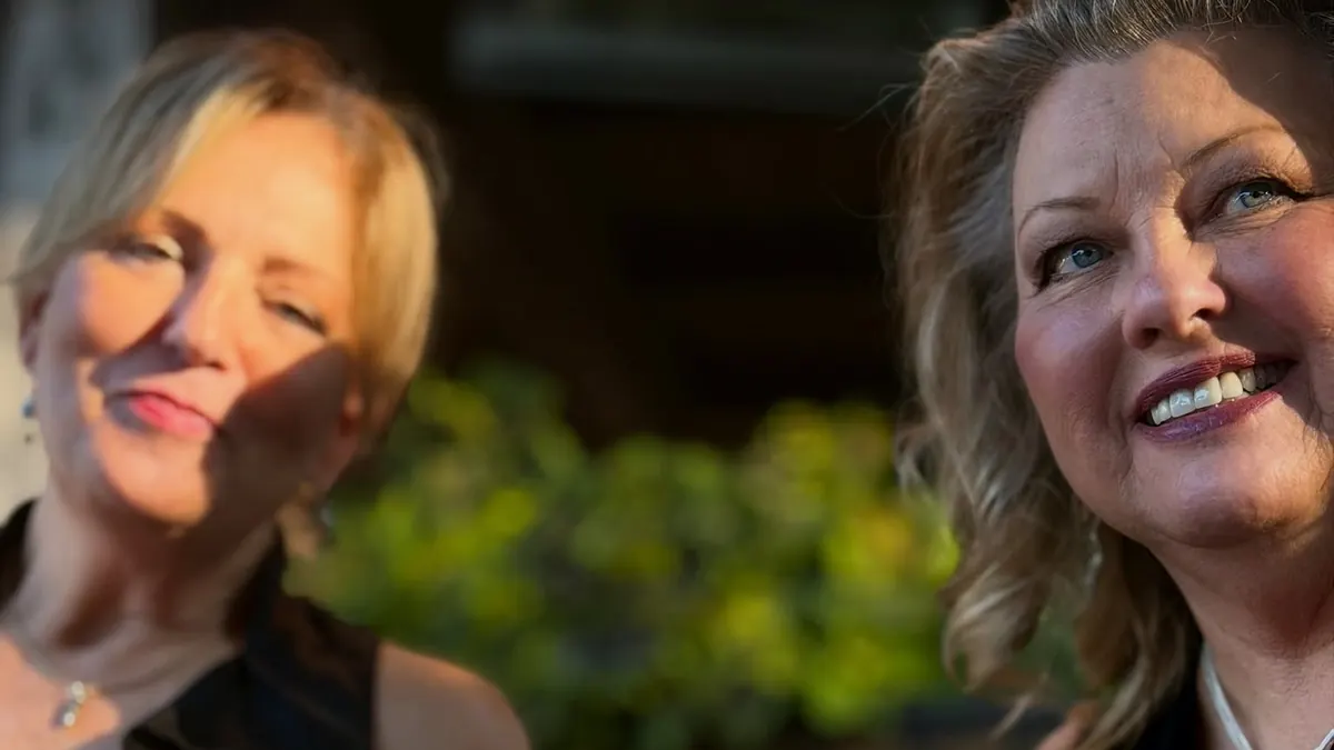 Two smiling women laughing together over cocktails and dessert at an outdoor table.