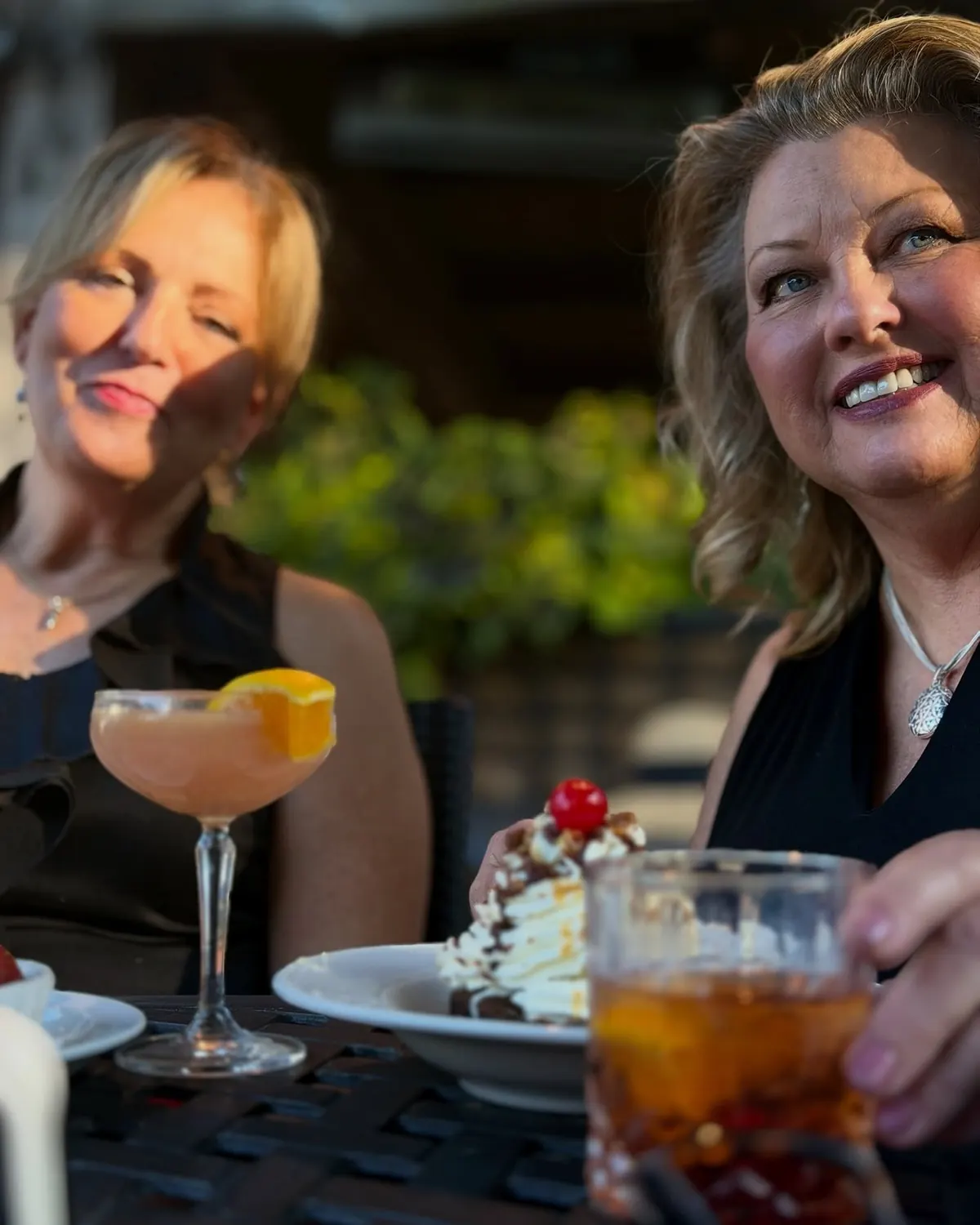 Two women laugh together over cocktails and dessert at an outdoor table.